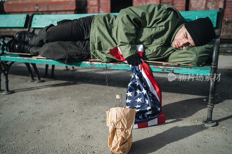无家可归的老兵拿着美国国旗和一瓶酒睡在长凳上