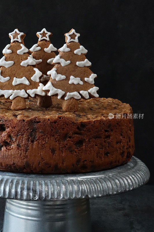 自制圣诞水果蛋糕的特写图像，顶部是用白色皇家糖霜覆盖的姜饼圣诞树饼干，放在银色的蛋糕架上，黑色的背景，前景聚焦