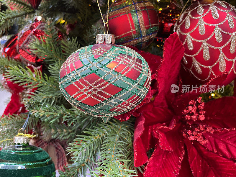 近距离图像的红色，绿色和金色闪光格子图案的装饰物挂在人工圣诞树的树枝上，人工的红色一品红，云杉针，模糊的白色神仙灯的背景
