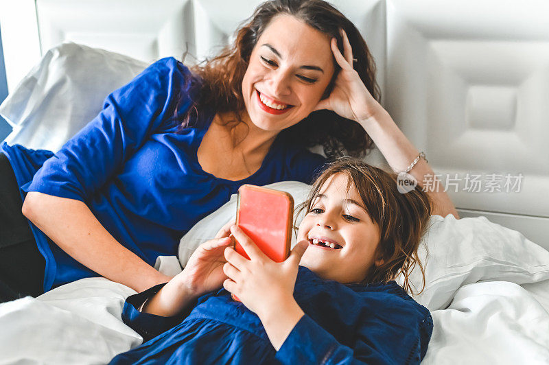 妈妈和女儿一起在床上放松地玩着，用手机看视频，拥有科技的幸福家庭