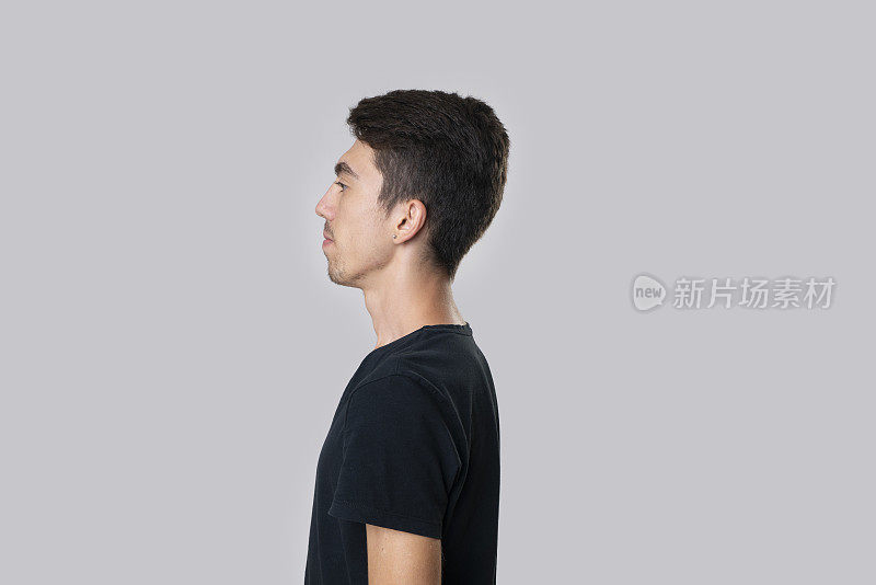 一个穿着黑色t恤的年轻男子的肖像，左转，侧视图，面无表情，。摄影棚拍摄，灰色背景。