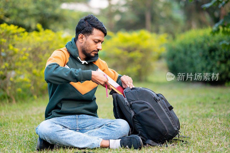 年轻的学生坐在公园里从背包里拿书。