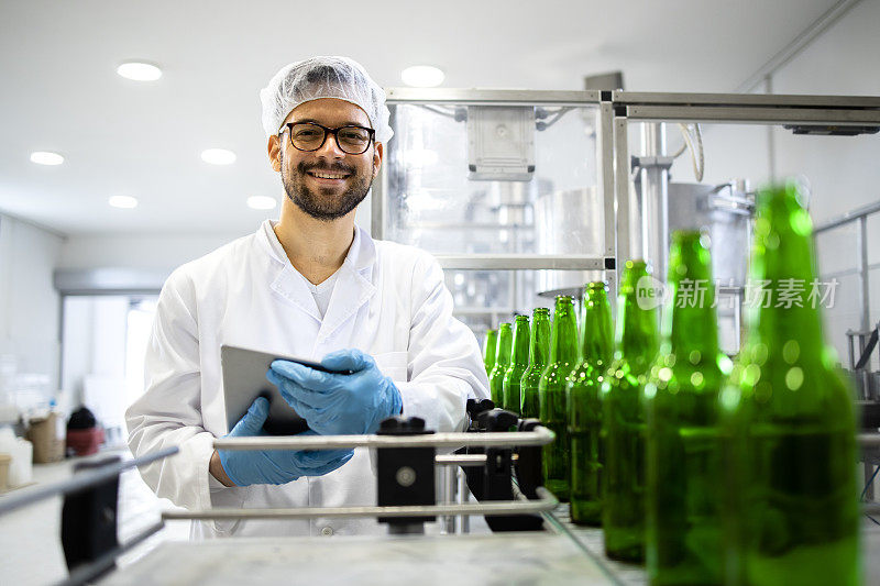 站在啤酒装瓶厂检查产品质量的经验丰富的专业技术人员的肖像。在后台生产线机器和啤酒瓶在一起。