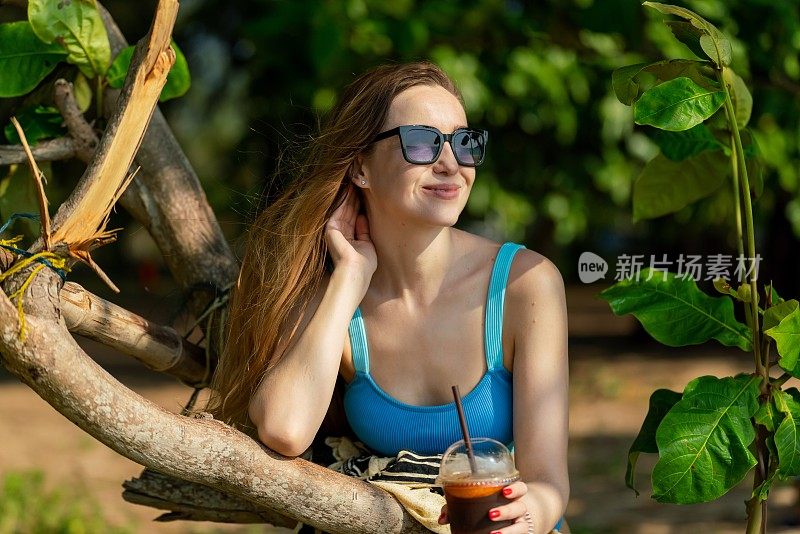 美丽的白人女性戴着太阳镜和蓝色比基尼，在海滩旁郁郁葱葱的热带树木公园里喝着混合冰饮料