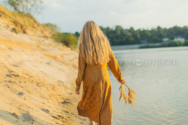 后面的观点。穿黄色衣服的金发女人躺在河岸上。夏天的一天。长头发的。沙子。