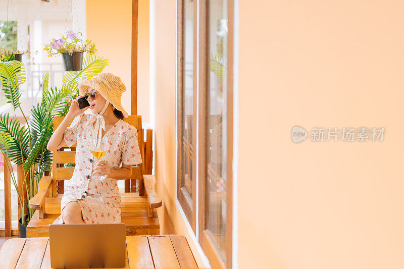 一名亚洲女性游客穿着裙子，戴着眼镜和帽子，坐在房间前的电话旁，喝着一杯清爽的葡萄酒。桌子上有一台笔记本电脑。