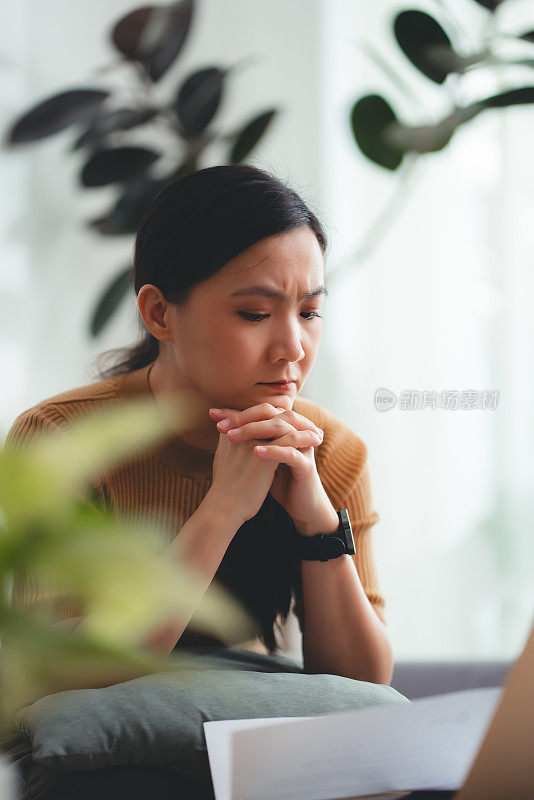 亚洲女性坐在沙发上，担心银行票据、抵押贷款债务和生活成本的账单支付问题。