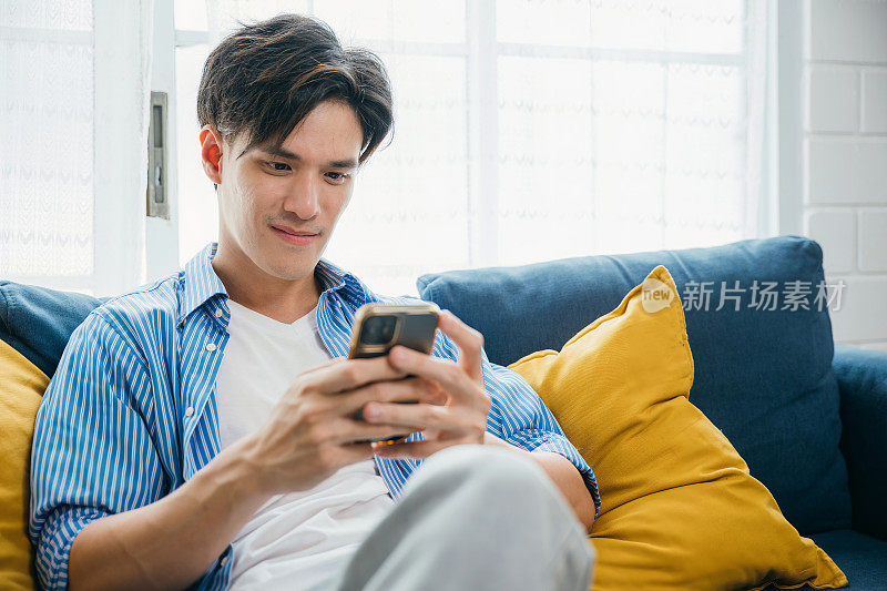 在家里的沙发上，一个面带微笑的年轻人在打电话，拥抱通讯和科技。他的幸福反映了成功的生活和放松。刷社交媒体。
