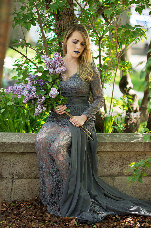 年轻的金发女人穿着灰色的蕾丝连衣裙，坐在长满紫丁香的花园墙上。