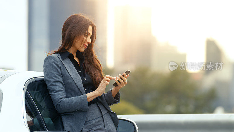 商务女性靠在高级轿车上使用智能手机。以摩天大楼为背景的大城市
