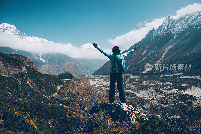 站在山上的年轻女子背着背包，举起双臂，望着群山。风景与快乐的女孩，高山，蓝天与云朵在秋天的尼泊尔。旅行。徒步旅行在喜马拉雅山脉