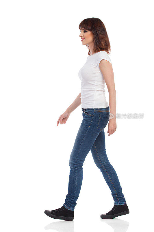 穿着牛仔裤和白色t恤的年轻女人正在走路。侧视图。