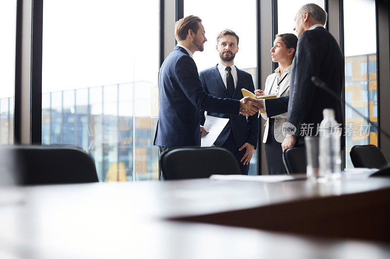 在会议中结识新的业务伙伴:内容满意的年轻的大胡子商人与成熟的总监握手，并在商业论坛后与同事交谈