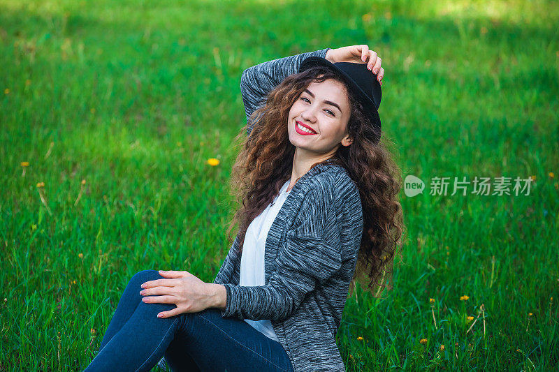 有魅力的女孩戴着帽子坐在绿色的草地上。