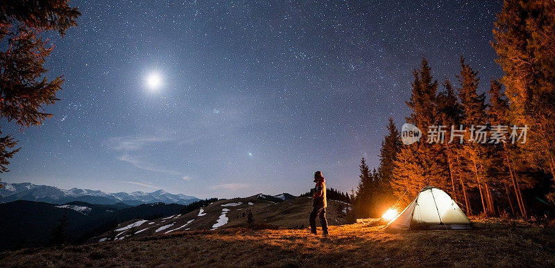 男游客晚上在森林附近的营地休息。在满是星星和月亮的美丽夜空下，一个人站在篝火和帐篷旁，欣赏着夜景。全景景观