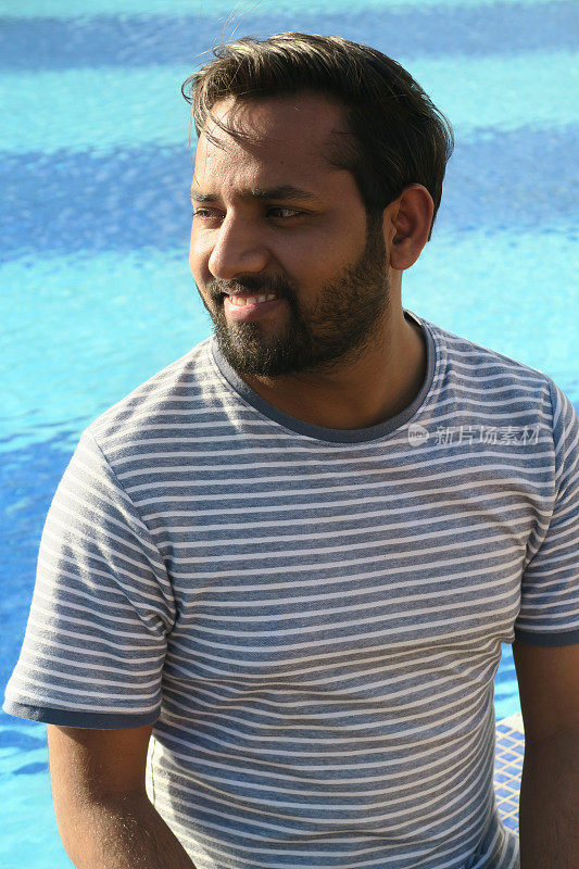 英俊的年轻印度男子坐在游泳池在阳光下看起来快乐的暑假，牙齿微笑，大笑，好看的印度男子享受阳光无限游泳池蓝色马赛克瓷砖，度假模式在牛仔夹克