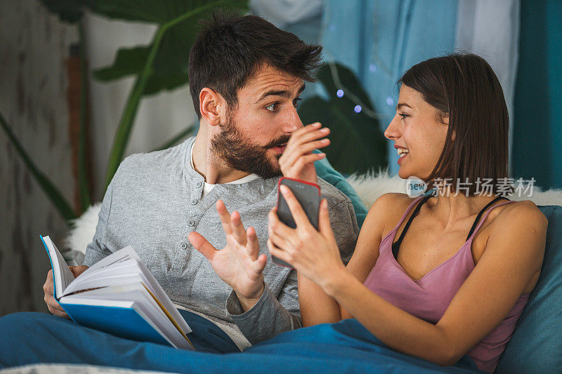 年轻夫妇在床上看书，使用智能手机，并有一个轻松的谈话