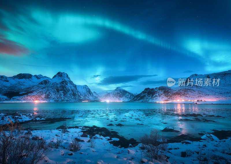 北极光越过海岸，雪山和城市的灯光在晚上。挪威罗浮敦群岛的北极光。星空与极地光。冬季景观与极光反射在水中