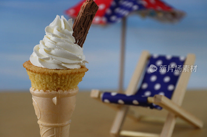 近距离的冰淇淋蛋筒设计纸杯蛋糕站在折叠椅和遮阳伞前，自制香草蛋糕上面覆盖着糖霜和巧克力薄片，海滩场景的背景，聚焦前景