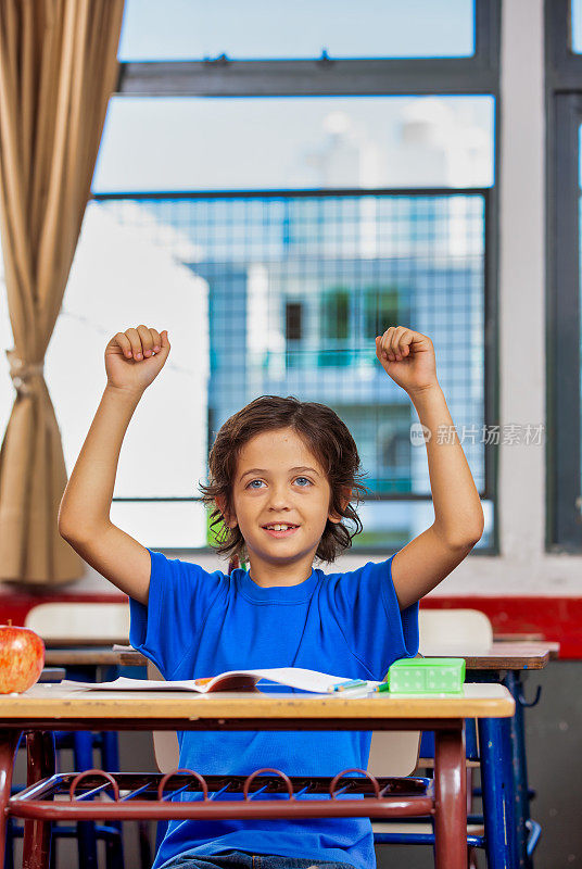 回到学校。教育的概念。快乐的阿根廷男学生坐在书桌前举起双手庆祝胜利。开学第一天