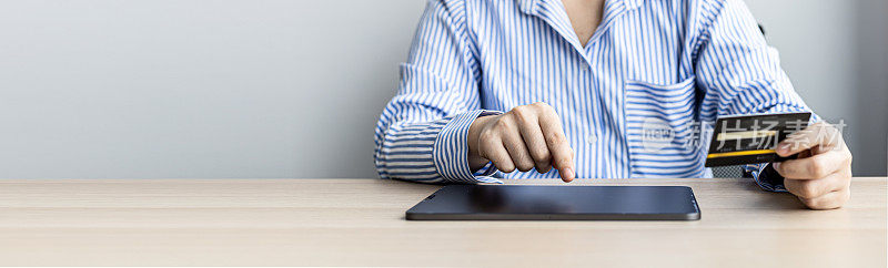 一位女士手持信用卡在平板电脑上打字，她在笔记本电脑上的购物应用程序中填写信用卡信息，为订单付款。使用信用卡支付的网上购物概念。