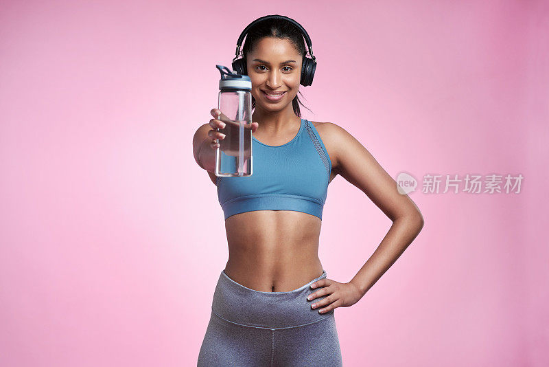 裁剪的肖像，一个迷人的和运动的年轻女子戴着耳机和摆姿势与一个水瓶在工作室的粉红色背景