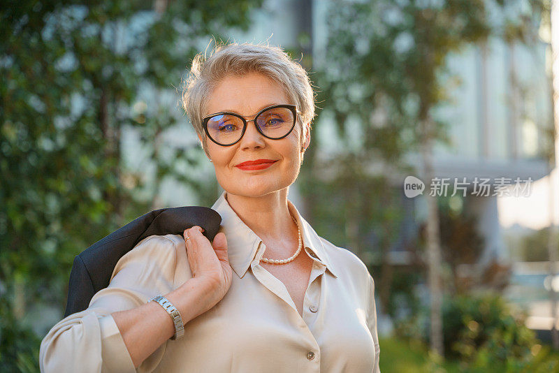 在一个夏日的傍晚，站在街道上的一位头发花白、戴着眼镜、留着短发、面带微笑的女商人站在镜头前