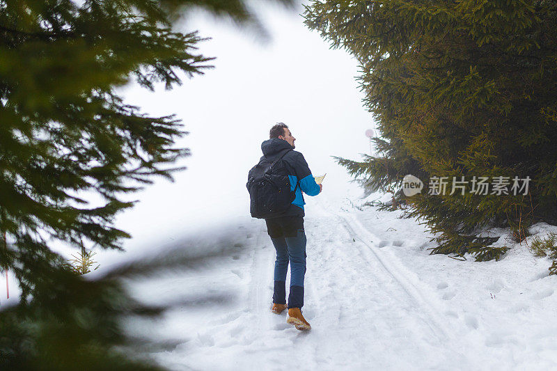 年轻的男性徒步旅行者穿着蓝色的夹克，在雾蒙蒙的雪山上走着，手里拿着地图