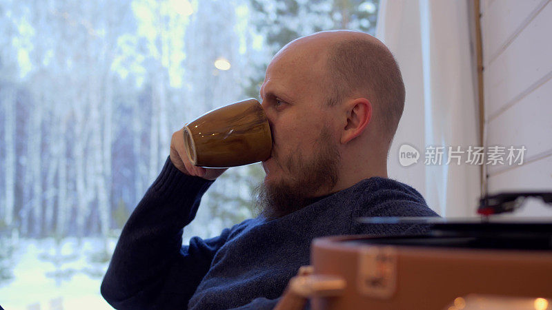 一个留着胡子的中年男子坐在家里，用一个大杯子喝咖啡。一个男人在听黑胶唱片留声机里的音乐。男人失去后的孤独。