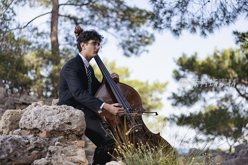 身穿黑色西装的年轻音乐家在大自然中演奏低音提琴。
