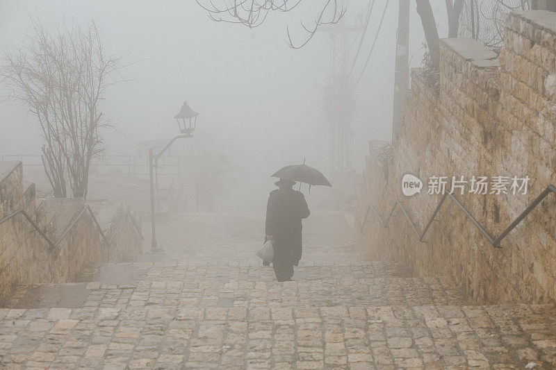 一位戴着帽子、穿着外套的老人打着一把黑伞，在雾中走上楼梯。孤独悲伤的男人打着雨伞在雨中行走。一个老人的背影。悲伤,思念,孤独