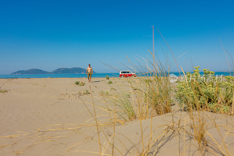光着膀子的男人走在沙滩上，露营车停在背景