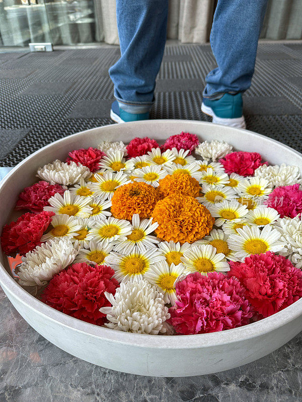 印度乌拉里碗的图片，里面装满了粉红色、橙色和白色的花朵——雏菊、万金菊和菊花漂浮在一盘水中，圆形的插花展示，腿在背景中，高架视图