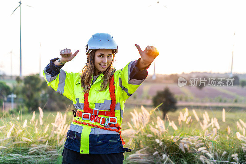 在风力涡轮机农场，穿着制服的白人女工程师微笑着竖起大拇指，庆祝项目成功。快乐工作者享受工作
