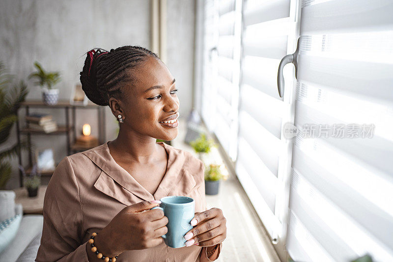 微笑的黑人妇女站在窗边喝咖啡，向外看