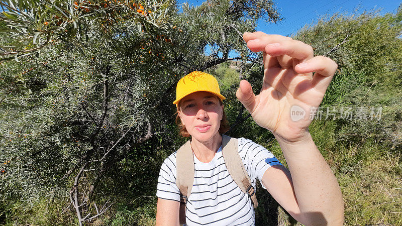 一位成熟的女性游客在小山城的河岸边品尝沙棘果。自拍视角