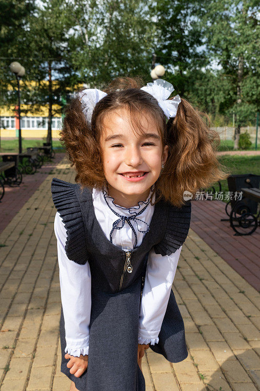 在学校操场上穿着系着白色蝴蝶结的校服，笑容无牙的开朗有趣的女孩。9月1日返校。一个快乐的小学生。小学教育，小学课堂。学生肖像