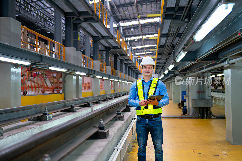 专业的工程师工人拿着平板电脑，微笑着看着相机，站在工厂工作场所的铁轨旁。