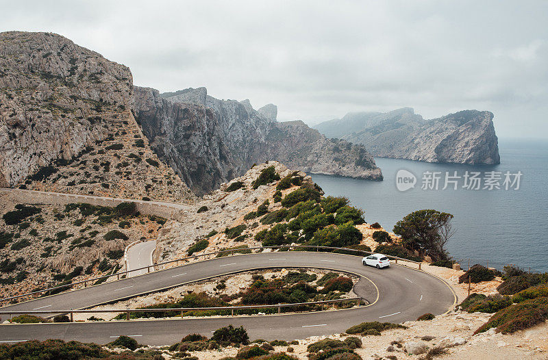 孤独的小白车行驶在弯弯曲曲的沥青山路附近，福门托角灯塔附近有美丽的海景和岩石海岸。西班牙巴利阿里群岛的马略卡岛。