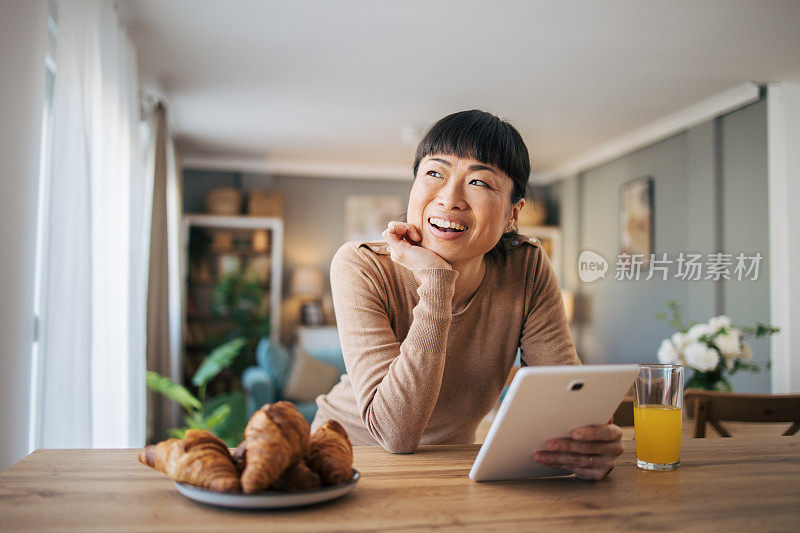 一名日本中年女性穿着睡衣坐在家里查看社交媒体或在她的数码平板电脑上远程工作。