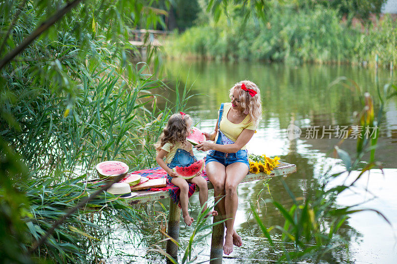 金发碧眼的母亲带着三岁的女儿，穿着牛仔短裤，系着红色的蝴蝶结，坐在码头的格子毯子上吃西瓜，河边有一束盛开的向日葵
