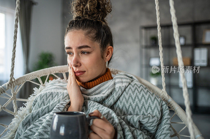 年轻女子喝着热茶或咖啡，感到牙痛，坐在家里客厅的秋千上。