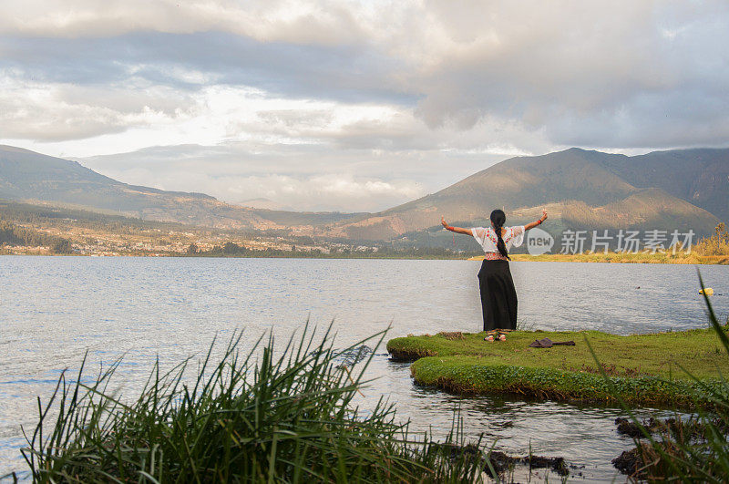 一位来自奥塔瓦洛(Otavalo)的土著妇女站在长满草的湖边，高举双臂，拥抱平静的早晨，同时进行放松和冥想练习。