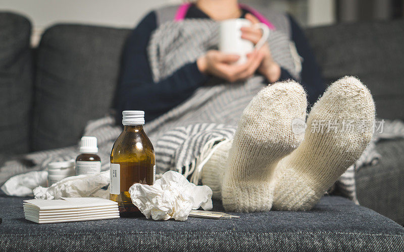 感冒药和生病的女人喝热饮来恢复流感，发烧和病毒。桌子上有脏的纸巾和纸巾。生病的人在冬天穿着暖和的羊毛长袜。