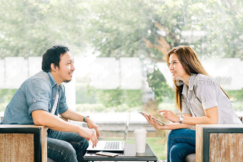 年轻的亚洲夫妇或同事在咖啡馆或现代的办公室里聊天，背景是花园。有笔记本电脑、智能手机和数字平板电脑。现代生活方式与电脑小配件技术概念
