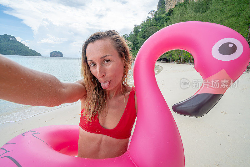 在泰国群岛，一名年轻女子在田园诗般的海滩上与粉色充气火烈鸟自拍。人们旅游目的地有趣和酷的态度概念