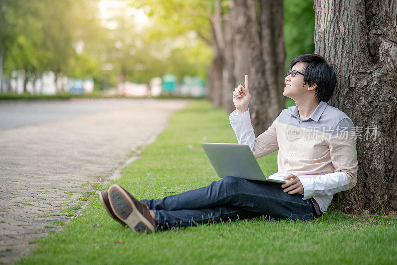 年轻的亚洲人大学学生戴着眼镜和笔记本电脑坐在绿色的草地上手指向上在大学公园。教育机会或奖学金。想法和灵感概念