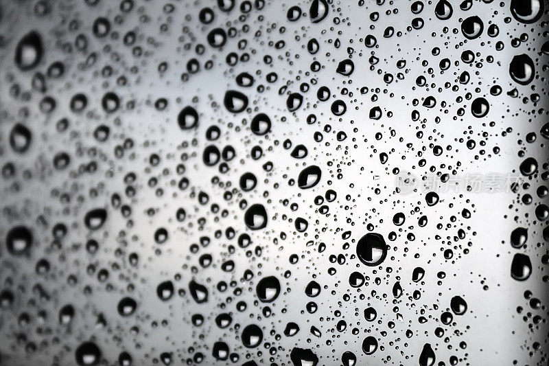 在一个雨天，玻璃上的黑色水滴带着白色的辉光，映衬着模糊多云的天空背景。有选择性的重点