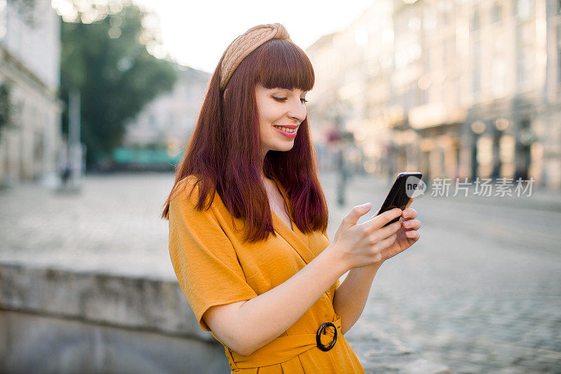 侧视图肖像的漂亮年轻的白人红头发的女人，穿着黄色工作服，站在外面的老城街道和手机短信。老城区有手机的女孩