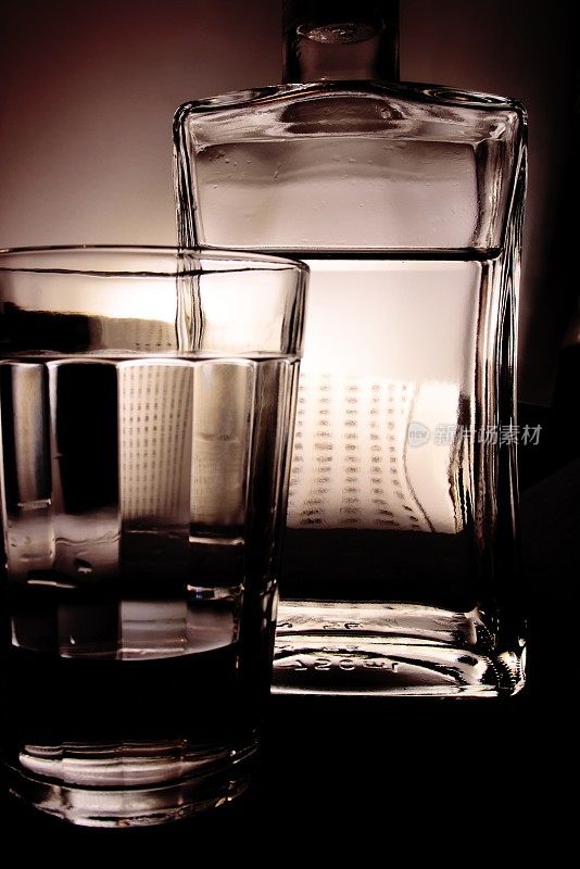 一个长方形的瓶子和一个透明的玻璃杯，装满了无色的液体，在半影的背景上。
